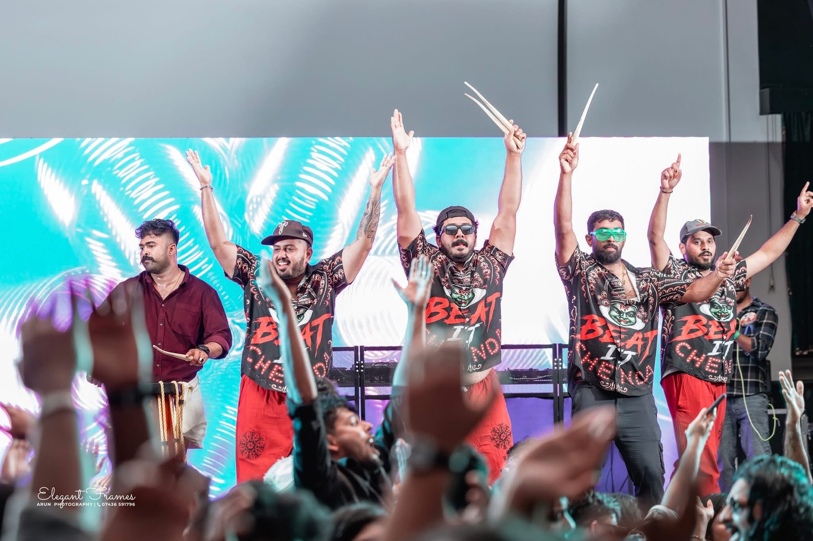 Chenda performers raising their drumsticks and hands against a turquoise stage backdrop